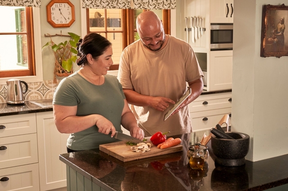 A man and woman cooking in their home kitchen, the man is pointing to a recipe on his tablet. 