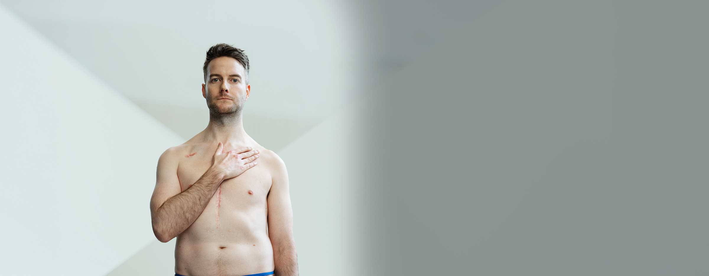 Geoff Lester shirtless with his hand on heart, visible scar from heart surgery