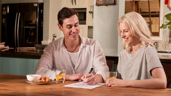A couple sitting at a wooden table in their kitchen, writing their Will. The man, wearing a white shirt, is holding a pen and writing on a piece of paper while the woman, in a gray t-shirt, looks on.